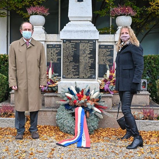 Zwei Personen stehen vor einem Gedenkstein, tragen Masken und Lederstiefel. Der Gedenkstein hat einen Kranz und ein Band mit den Farben Rot, Weiß und Blau.