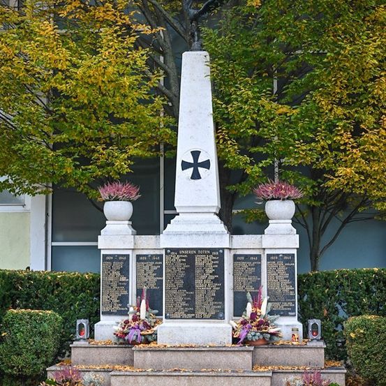 Ein Steinmonument mit einem Kreuz oben und Tafeln mit Namen, umgeben von Pflanzen und Hecken in einer Außenumgebung.