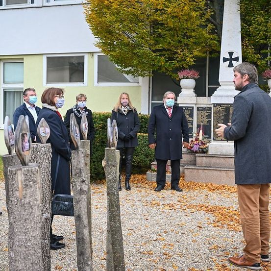 Eine Gruppe von Menschen in Winterkleidung steht vor einem Denkmal mit einem Kreuz und diskutiert, während ein Mann vor ihnen spricht.
