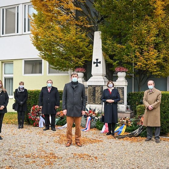 Eine Gruppe von Menschen in Wintermänteln und Mundmasken steht vor einem Denkmal mit einem Kreuz und Tafeln, umgeben von Blumen und Herbstblättern.