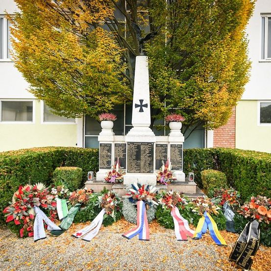Ein Denkmal mit einem Kreuz oben, umgeben von Blumen und Bändern, vor einem Gebäude mit gelben Blättern.