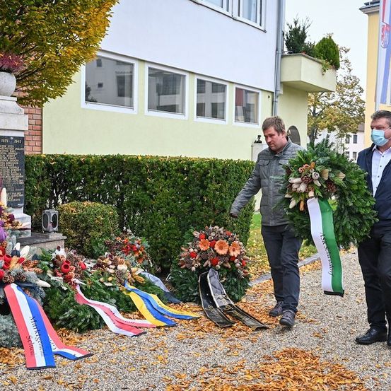 Zwei Männer in formeller Kleidung platzieren Kränze an einer Gedenkstätte. Das Denkmal ist mit Blumen und Bändern geschmückt. Hinter ihnen ist ein Gebäude mit Glasfenstern und einer Flagge zu sehen.