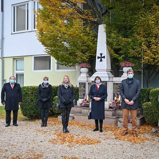 Eine Gruppe von Menschen mit Masken steht vor einem Denkmal in einem Park im Herbst.