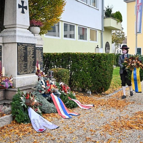 Zwei Personen in traditioneller Kleidung platzieren Kränze an einem Denkmal. Das Denkmal hat ein Kreuz und eine Tafel mit Namen. Die Szene spielt sich in einem Park mit Büschen und einem Gebäude im Hintergrund ab.
