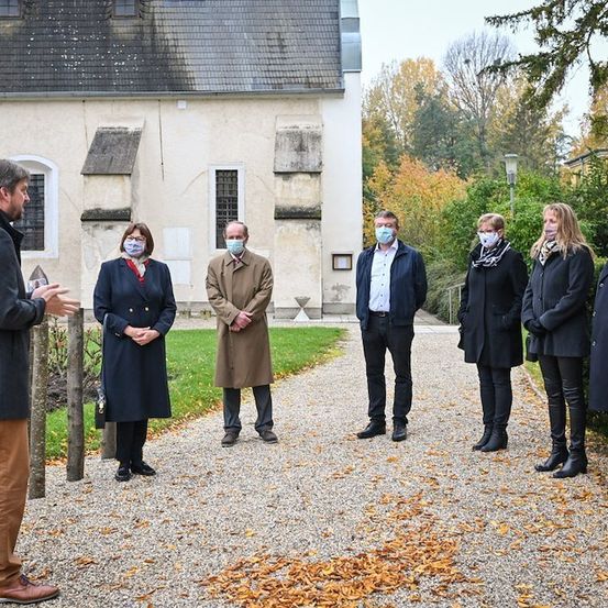 Eine Gruppe von Menschen in Winterkleidung, einschließlich Masken, steht vor einem historischen Gebäude mit Herbstlaub im Hintergrund.