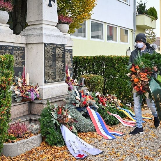 Ein Mann mit Maske und Mütze legt einen Kranz auf ein Denkmal mit einer Tafel und anderen Kränzen auf dem Boden.