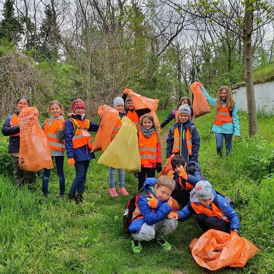 Eine Gruppe von Kindern in Warnwesten sammelt Müll in einem Grasbereich. Sie halten orange und gelbe Müllsäcke.