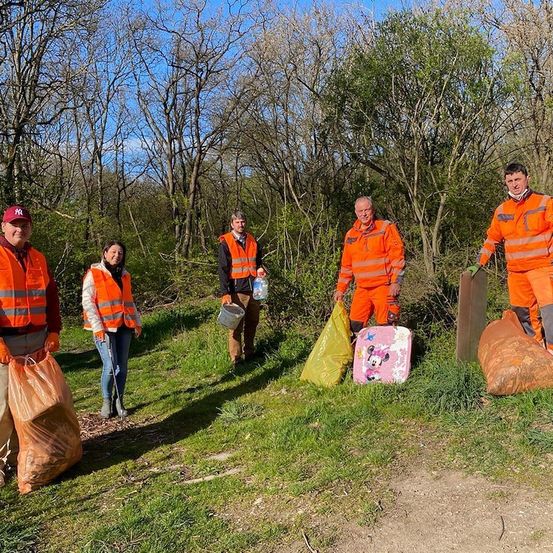 Fünf Personen in orangefarbenen Westen stehen in einem Grasbereich, möglicherweise an einer Reinigungsveranstaltung teilnehmend. Sie halten verschiedene Gegenstände, darunter einen Eimer, einen Beutel und einen Plastikbeutel. Das Gebiet ist von Bäumen und Büschen umgeben.