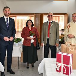 Vier Personen in formeller Kleidung stehen in einem Raum mit einem Tisch, auf dem sich ein Geschenkkarton und eine Pflanze befinden. Hinter ihnen ist ein Fenster mit Jalousien und eine Tür zu sehen.