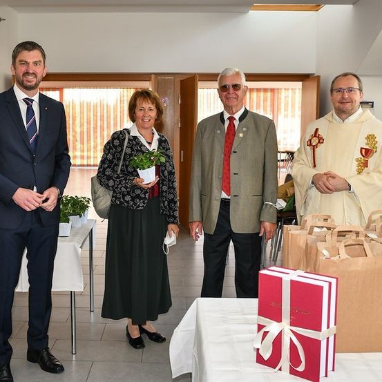 Vier Personen stehen in einem Raum, zwei Männer und zwei Frauen, eine Frau hält eine Topfpflanze. Ein Tisch mit einem Geschenkkarton und Papiertüten steht vor ihnen.