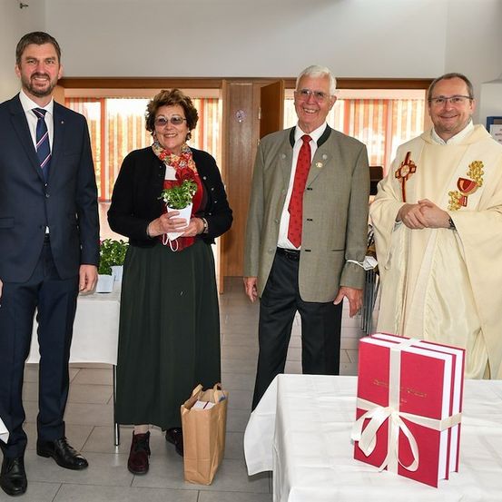 Vier Personen stehen in einem Raum zusammen, einer hält eine Topfpflanze, ein anderer einen Papierbeutel, und zwei Männer in formeller Kleidung. Ein Priester steht neben einem Tisch mit einem Geschenkkarton.
