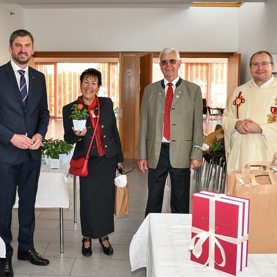 Vier Personen stehen in einem Raum, zwei Männer in Anzügen, eine Frau mit einer Blume und ein Priester. Sie lächeln und scheinen für ein Foto zu posieren. Ein Tisch mit einem Geschenkkarton und Taschen ist im Vordergrund.