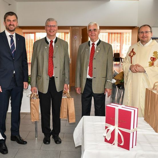 Vier Männer in formeller Kleidung, zwei mit Papiertüten, stehen um einen Tisch mit einer roten Geschenkbox und einem Priester. Die Umgebung scheint eine formelle Veranstaltung oder Zeremonie zu sein.