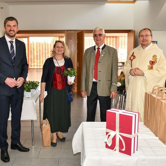 Vier Personen stehen um einen Tisch mit einem Geschenkkarton und einer Pflanze. Der Mann links trägt einen Anzug und eine Krawatte, während die Frau in der Mitte eine Topfpflanze hält. Der Mann in der Mitte trägt eine rote Krawatte und eine Jacke. Der Mann rechts trägt eine weiße Robe.
