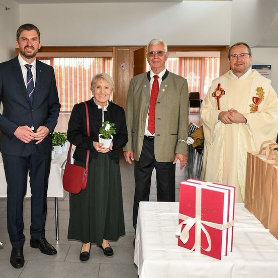 Vier Personen stehen in einem Raum, zwei Männer in Anzügen, eine Frau mit einem Topfpflanzen und ein Priester mit einem roten und weißen Gewand. Ein Tisch mit einem Geschenkkarton und Taschen steht vor ihnen.