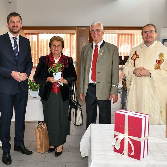 Vier Personen stehen in einem Raum, zwei Männer in Anzügen, eine Frau mit einer Blume und ein Priester in traditioneller Kleidung. Ein Tisch vor ihnen hält eine Geschenkbox.