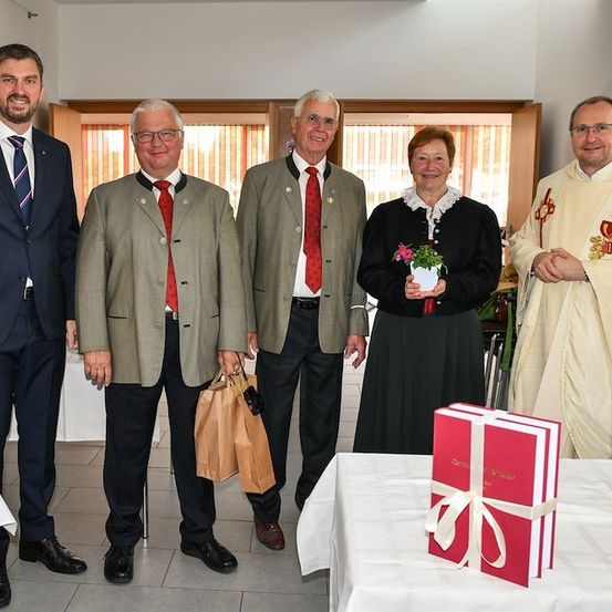Fünf Personen in formeller Kleidung stehen um einen Tisch mit einem Geschenkkarton, eine Frau hält einen Blumentopf. Der Mann rechts trägt eine Priesterrobe.