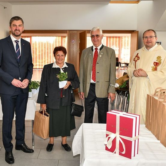 Vier Menschen stehen in einem Raum, zwei Männer und zwei Frauen, lächeln für ein Foto. Die Frau in der Mitte hält eine Topfpflanze und eine Handtasche. Der Mann auf der linken Seite trägt einen Anzug und eine Krawatte. Der Mann auf der rechten Seite trägt eine Soutane. Ein Tisch mit einem Geschenkkarton und Papiertüten steht vor ihnen.