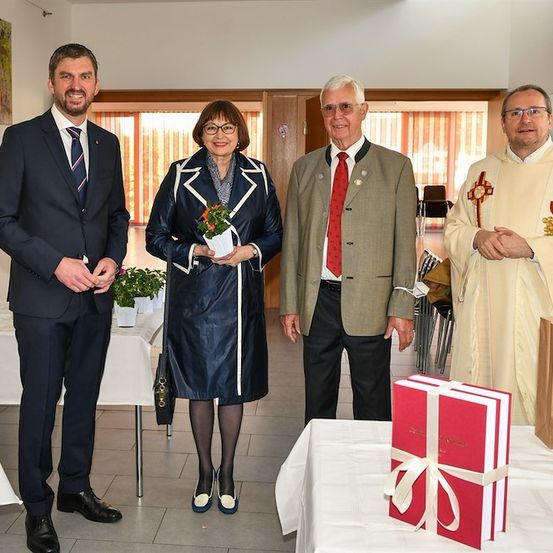 Vier formell gekleidete Personen stehen in einem Raum, wobei eine Frau eine Blume hält, ein Mann mit roter Krawatte und ein Priester im Hintergrund. Ein Tisch mit einem Geschenkkarton steht im Vordergrund.