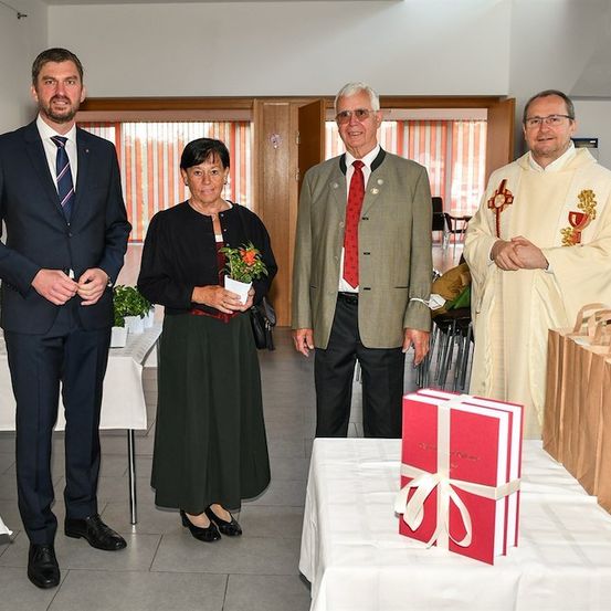 Vier Personen stehen um einen Tisch mit Geschenkbox und Papiertüten. Ein Mann in Anzug hält eine Blume, während eine Frau in schwarzem Kleid eine Topfpflanze hält. Ein weiterer Mann in grauem Anzug und ein Priester in weißer Robe stehen neben ihnen.