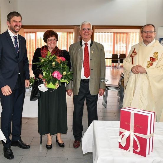 Vier Personen stehen in einem Raum mit einem Priester, der einen Blumenstrauß hält, einem Mann in einem Anzug, der ein Buch hält, und zwei weiteren Männern in Anzügen und Krawatten.