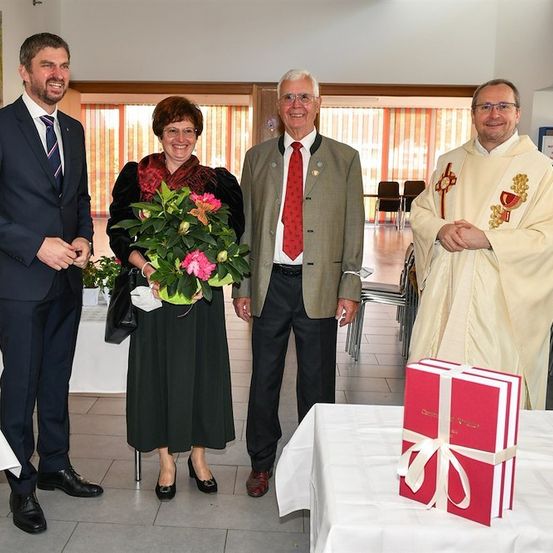 Vier Personen stehen in einem Raum, lächeln und posieren für ein Foto. Eine Frau hält einen Blumenstrauß, und ein Priester steht neben einer Geschenkbox.