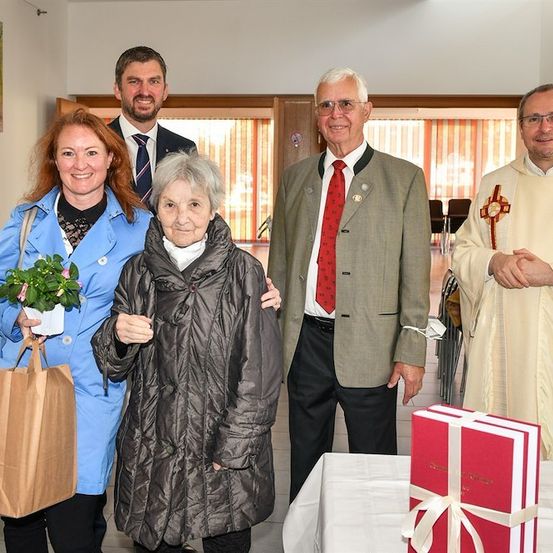 Eine Gruppe von Menschen, einschließlich eines Priesters, steht lächelnd in einem Raum zusammen, um ein Foto zu machen. Eine Frau hält eine Topfpflanze und einen Papierbeutel. Ein Mann im Anzug hält den Arm einer Frau. Ein Tisch mit einem Geschenkkasten steht vor ihnen.