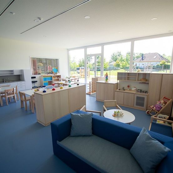 Ein gut beleuchteter Indoor-Spielbereich für Kinder mit Holzmöbeln, einer blauen Couch und großen Fenstern, die einen sonnigen Außenspielplatz zeigen.