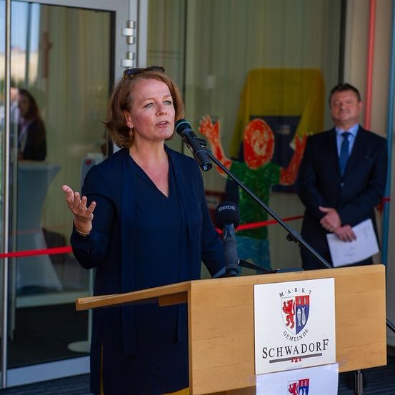 Eine Frau spricht an einem Podium mit Mikrofon, in einem blauen Blazer. Hinter ihr steht ein Mann mit einem Papier, und ein bunter Banner ist sichtbar. Der Hintergrund umfasst eine Glastür und eine gelbe Tischdecke.