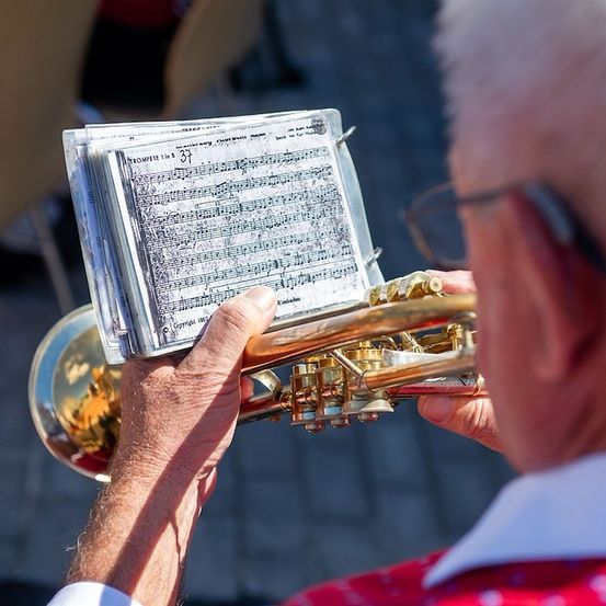 Ein älterer Mann spielt eine Trompete und hält ein Notenblatt in den Händen. Er trägt eine Brille und ein rotes Hemd.