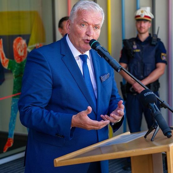 Ein Mann in einem blauen Anzug spricht in ein Mikrofon an einem Podium, mit einem Polizeibeamten hinter ihm.