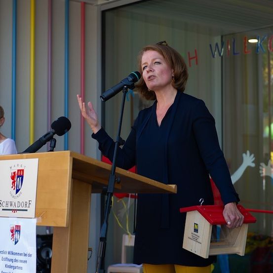 Eine Frau steht an einem Rednerpult und spricht in ein Mikrofon, mit einem Schild, das 'SCHWADORE' und einem Banner, das 'Willkommen bei der Eröffnung des neuen Kindergartens' sagt. Hinter ihr zeigt eine Glaswand bunte Buchstaben und einen Baum.