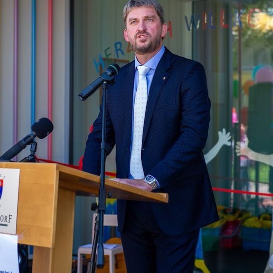 Ein Mann in einem Anzug steht an einem Podium mit einem Mikrofon, möglicherweise hält er eine Rede. Hinter ihm befindet sich eine Glaswand mit bunten Zeichnungen und dem Wort 'Herz'.