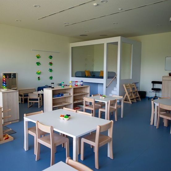 Ein gut organisierter Spielraum mit weißen Wänden und blauem Boden. Er verfügt über verschiedene Holzmöbel, einschließlich Tische, Stühle und Regale. Es gibt ein kleines Bett mit Kissen und ein weißes Spielhaus mit einer Treppe.