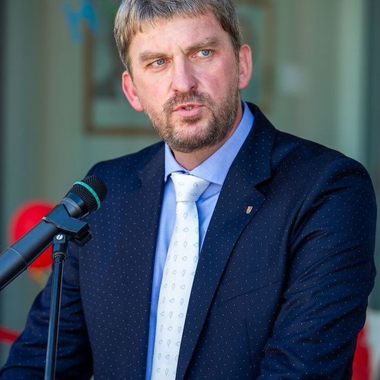 Ein Mann in einem blauen Anzug mit einer weißen Krawatte steht an einem Rednerpult mit einem Mikrofon und sieht ernst aus.