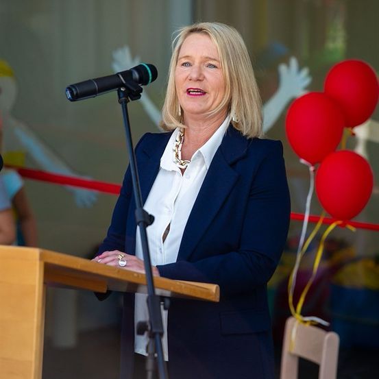 Eine Frau in einem blauen Blazer und einem weißen Hemd steht an einem Rednerpult und spricht in ein Mikrofon, mit drei roten Luftballons hinter ihr.