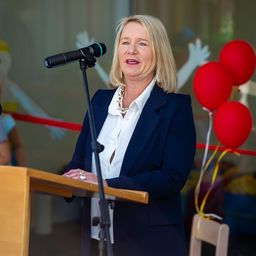Eine Frau in einem blauen Blazer und einem weißen Hemd steht an einem Rednerpult und spricht in ein Mikrofon, mit drei roten Luftballons hinter ihr.