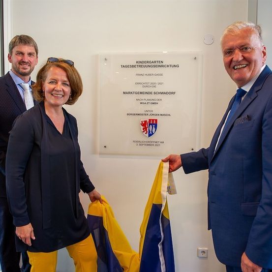 Drei Personen in formeller Kleidung stehen vor einer weißen Plakette mit Text und Logo, lächeln und halten eine Flagge.