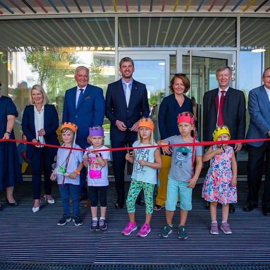 Eine Gruppe von Erwachsenen und Kindern steht vor einem Gebäude und hält ein rotes Band. Die Kinder tragen Kronen und halten Süßigkeiten. Die Erwachsenen tragen formelle Kleidung.