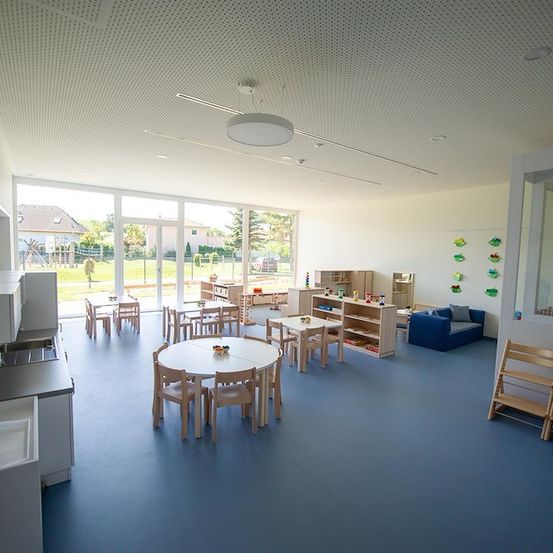 Ein leerer Klassenraum mit blauen Böden, weißen Wänden und großen Fenstern. Tische und Stühle sind ordentlich angeordnet. Ein blaues Sofa steht in der Nähe der Wand.