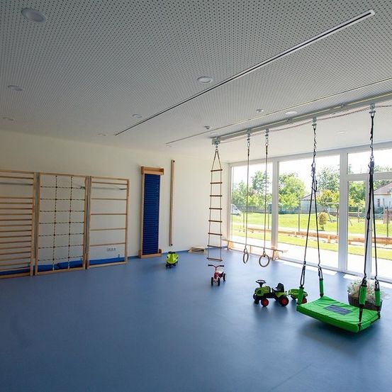 Ein Indoor-Spielbereich mit blauem Boden, großen Fenstern und verschiedenen Spielgeräten wie Schaukeln, Leitern und Spielzeug.