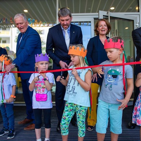 Eine Gruppe von Kindern und Erwachsenen versammelt sich für eine Eröffnungszeremonie. Die Kinder tragen Kronen und halten Scheren, während die Erwachsenen, einschließlich eines Mannes in einem blauen Anzug, hinter ihnen stehen. Die Szene spielt sich drinnen ab, mit einer Glaswand und einem Schild im Hintergrund.