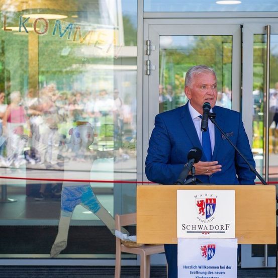 Ein Mann in einem blauen Anzug steht an einem Podium mit einem Mikrofon und spricht zu einem Publikum. Das Podium hat ein Schild mit der Aufschrift 'Marktgemeinde Schwadorf'. Im Hintergrund befindet sich eine Glaswand mit Reflexionen von Menschen und Bäumen.