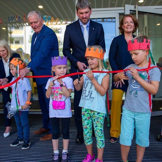 Eine Gruppe von Kindern und Erwachsenen versammelt sich für eine Eröffnungszeremonie. Die Kinder tragen Kronen und halten Scheren, während Erwachsene hinter ihnen stehen. Die Veranstaltung findet in einem modernen Gebäude mit Glastüren statt.