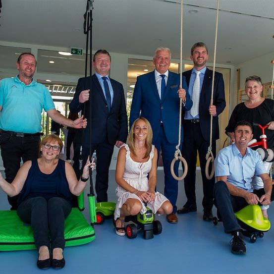 Eine Gruppe von Menschen in formeller Kleidung posiert für ein Foto in einem Raum mit blauem Boden. Einige sitzen auf grünen Schaukeln, während andere stehen. Eine Frau in einem weißen Kleid sitzt auf einer Schaukel und ein Mann in einem blauen Hemd sitzt auf einer grünen Schaukel. Eine Frau mit Brille sitzt auf einer Schaukel mit grüner Basis.
