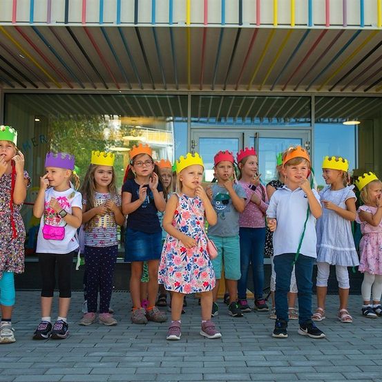 Eine Gruppe junger Kinder mit farbenfrohen Papierkronen steht in einer Reihe vor einem Gebäude mit einem mehrfarbigen Vordach.