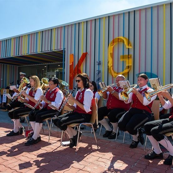 Eine Gruppe von Musikern in traditioneller Kleidung spielt auf Blechblasinstrumenten vor einem farbenfrohen Gebäude mit einem gelben Buchstaben G.