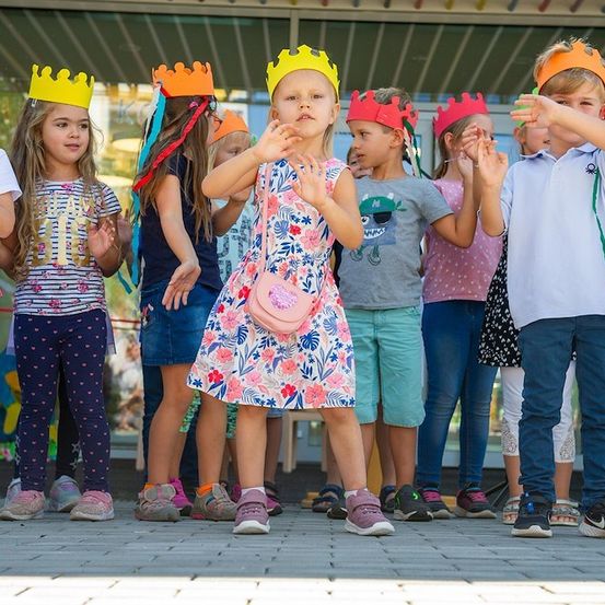 Eine Gruppe von Kindern mit Kronen, die in einer Reihe tanzen, einige mit erhobenen Händen, in einer Schulumgebung.
