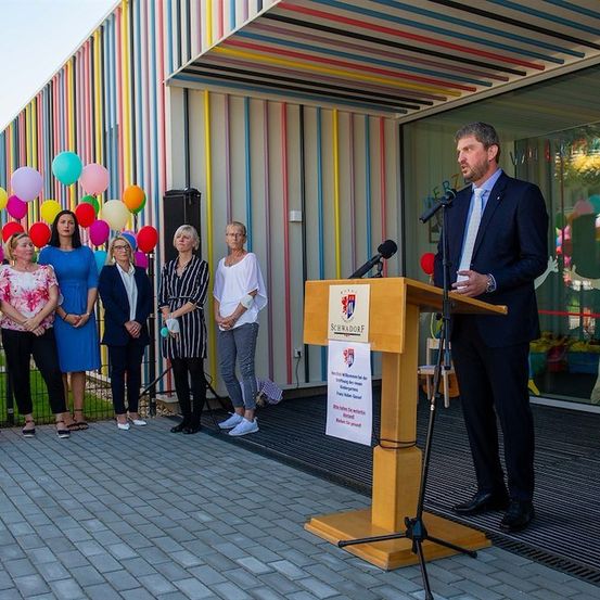Ein Mann in einem Anzug spricht an einem Podium mit einem Mikrofon, umgeben von einer Gruppe von Frauen vor einem farbenfrohen Gebäude mit Luftballons und einer Glaswand.