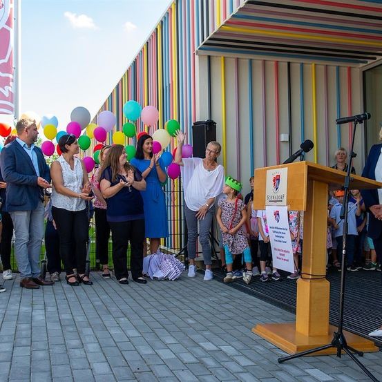 Eine Gruppe von Menschen, einschließlich Kindern, steht vor einem farbenfrohen Gebäude mit Luftballons, während eine Frau an einem Rednerpult spricht.
